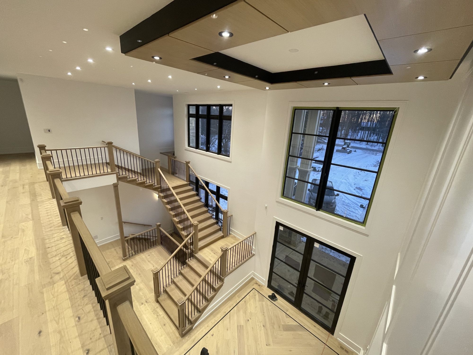 Two-storey open staircase in white oak with black spindles, an inset wood and black coffered ceiling, and a herringbone floor inlay