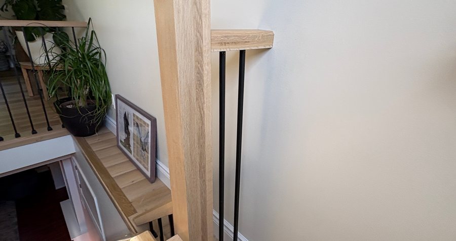 A custom oak stair rail and newel post with black metal balusters, joined against a painted wall.