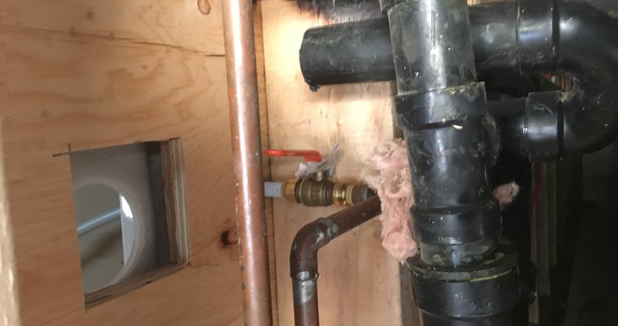 Copper and ABS plumbing rough-in behind an access panel, with supply valves labelled and insulation packed around the drain stack.