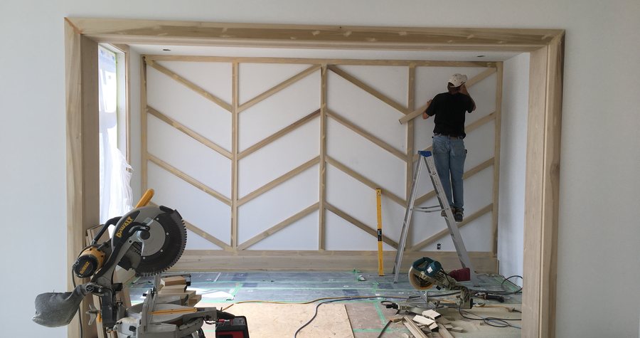 A trade installing a herringbone wood accent wall from a ladder, miter saw station set up in the room.