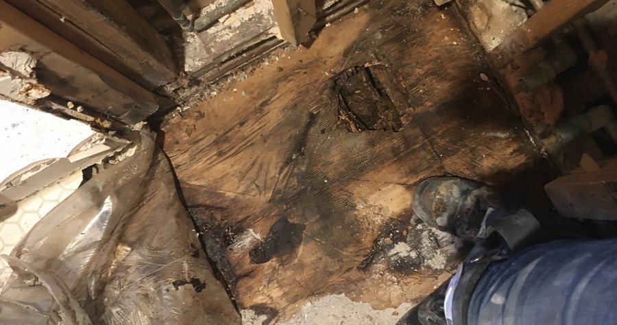 Subfloor rot exposed during a bathroom demolition, with old plumbing and framing visible and a worker's boot for scale.