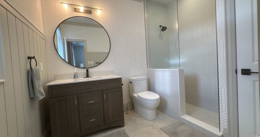 A finished Waterloo bathroom with wainscoting, geometric wallpaper, a round mirror and a curbless glass shower.